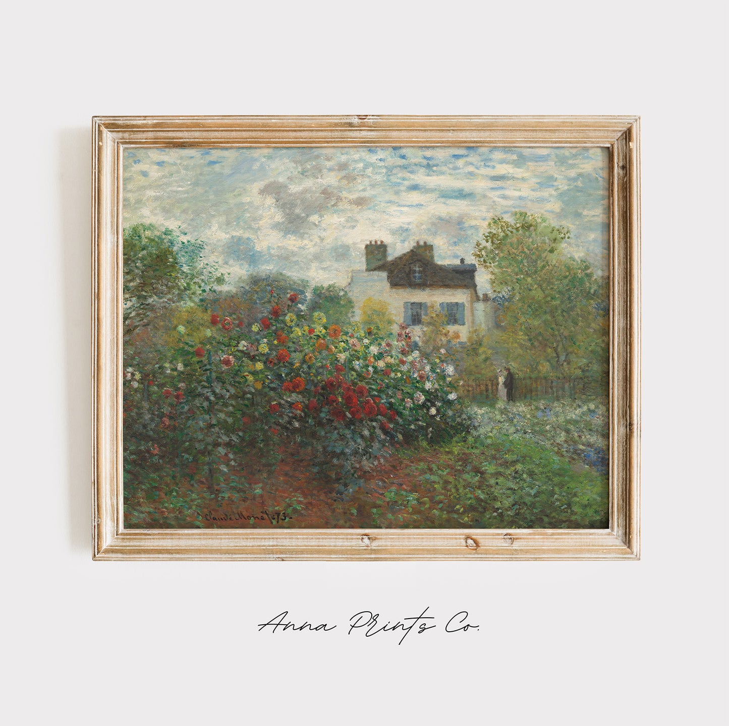 A framed Impressionist landscape painting featuring a white house surrounded by lush green trees and a vibrant red, pink, and white flower garden under a cloudy sky. The painting is housed in a light natural wood frame and is displayed against a clean white wall with 'Anna Prints Co' written in cursive below.