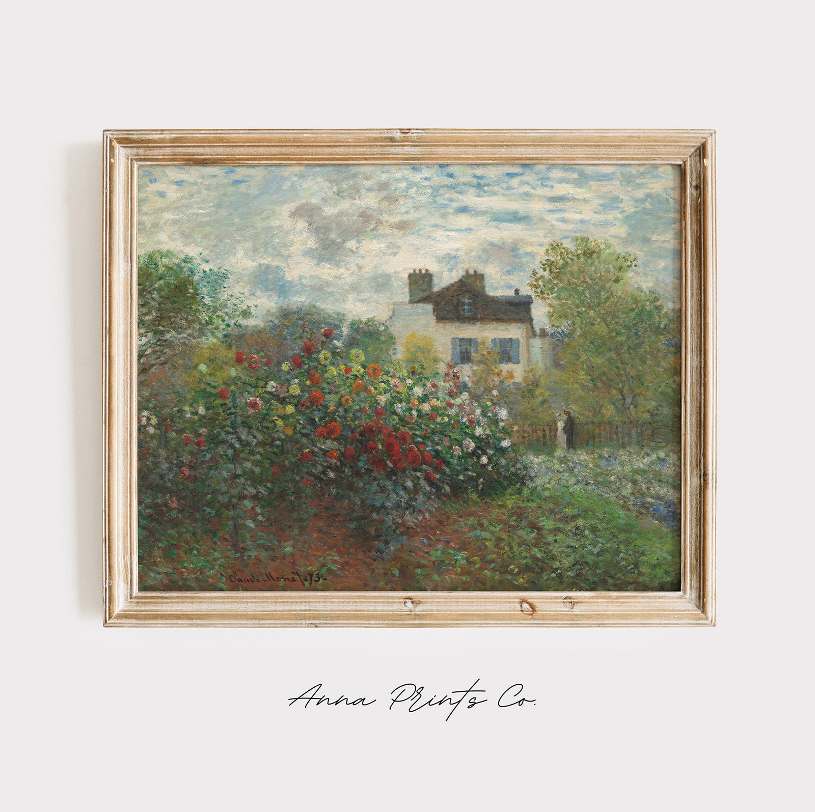 A framed Impressionist landscape painting featuring a white house surrounded by lush green trees and a vibrant red, pink, and white flower garden under a cloudy sky. The painting is housed in a light natural wood frame and is displayed against a clean white wall with 'Anna Prints Co' written in cursive below.