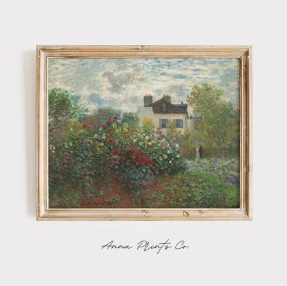 A framed Impressionist landscape painting featuring a white house surrounded by lush green trees and a vibrant red, pink, and white flower garden under a cloudy sky. The painting is housed in a light natural wood frame and is displayed against a clean white wall with 'Anna Prints Co' written in cursive below.